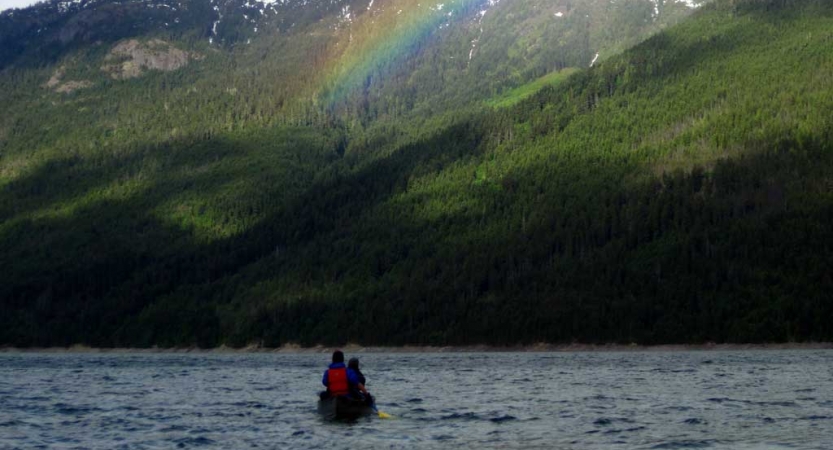 a canoe sits on a body of water facing a green mountain face with a rainbow cast in front of it